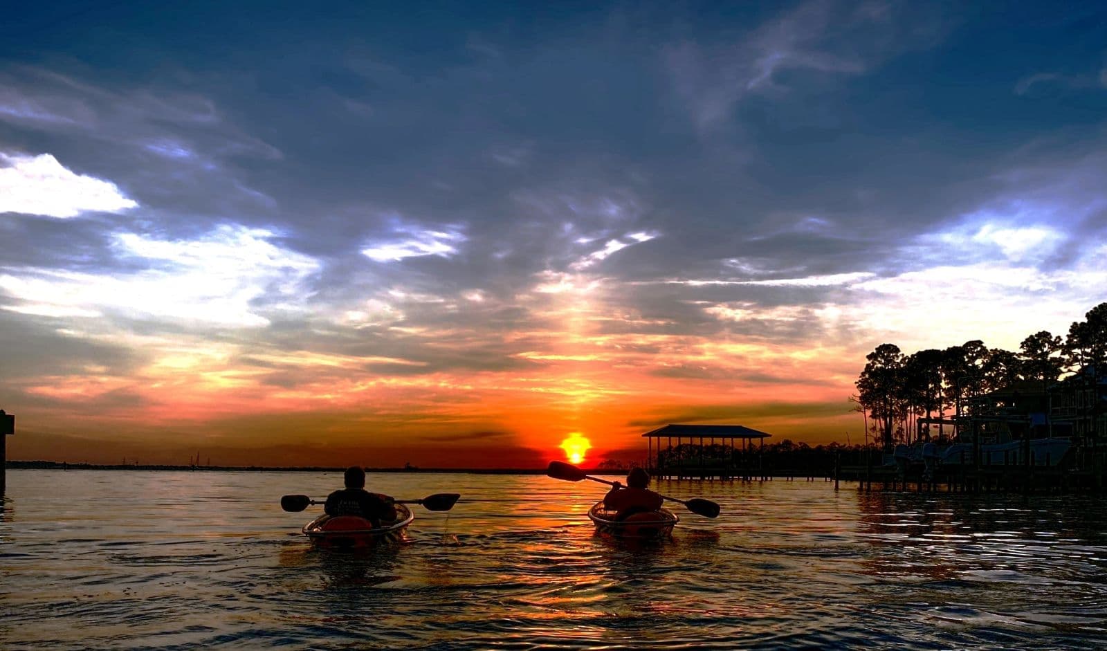 Two paddlers silhouetted against a dramatic orange sunset over Destin Harbor, dock and boathouse in the distance